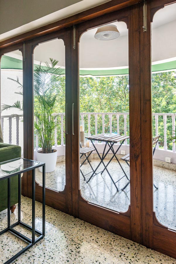 Big window with wooden frame looking over balcony with table and chairs set, lush green garden in the background