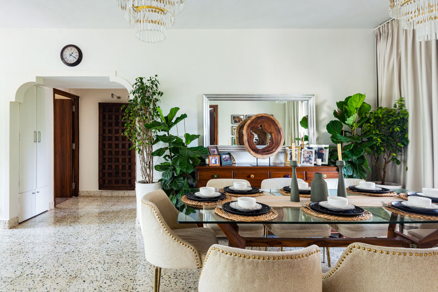 Bright living room with big dining table in glass and wood, wooden art piece and several big indoor plants