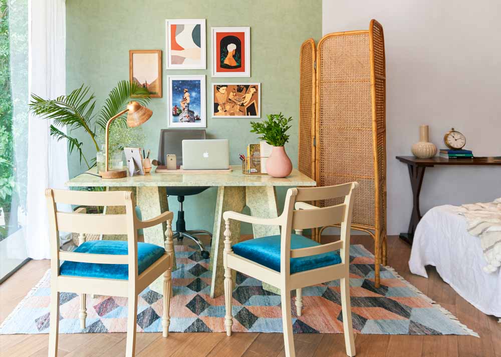 A room with a study table and two chairs, placed on a colourful rug, a rattan room divider and some frames hung on the wall